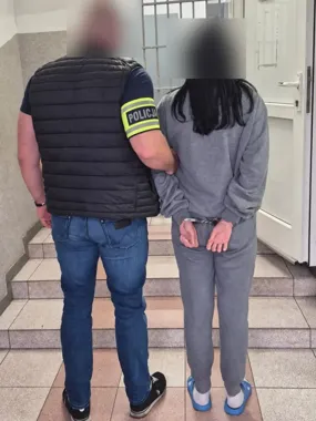 Funkcjonariusz policji w kamizelce z napisem "POLICJA" prowadzi zakutą w kajdanki kobietę w szarym dresie w budynku.