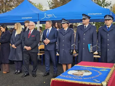 Grupa osób, w tym funkcjonariusze policji w mundurach, stoi pod niebieskimi namiotami z napisem „Legnica zachwyca” podczas uroczystości z udziałem sztandaru.