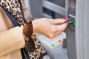 Kobieta w beżowym płaszczu i szalu w panterkę wkłada kartę do bankomatu.
