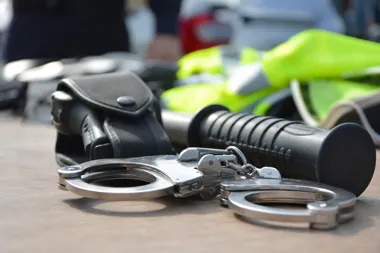 Policyjne kajdanki, pałka służbowa i kabura leżą na blacie, w tle widoczna odblaskowa kamizelka i samochód.